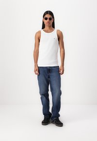 Weißes, geripptes Tanktop, blaue Denim-Jeans, schwarze Sneakers und runde Sonnenbrille. Auffällige Details: kleines Logo auf dem Tanktop und distressed Jeans.