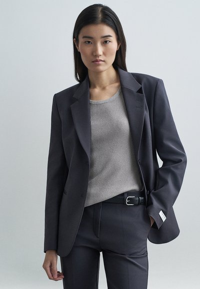 Donkergrijze op maat gemaakte blazer met notch lapels, twee knopen en subtiele stiksels, gedragen over een geribd lichtgrijs topje en broek.