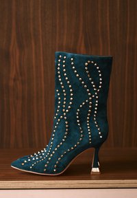 Cizmă de gleznă din piele întoarsă teal, cu ținte metalice aranjate în modele ondulate și toc subțire cu un accent auriu. Design cu vârf ascuțit.