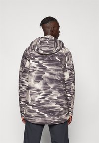 Chaqueta impermeable con capucha, con un patrón ondulado en blanco y negro. Está hecha de material sintético, tiene mangas largas y un dobladillo curvado.