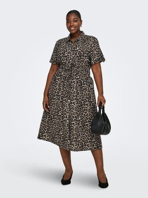 Femme debout, portant une robe léopard longueur genou et des talons noirs, tenant un sac à main noir dans sa main droite.