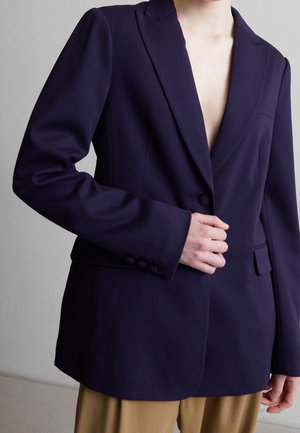 Osoba ubrana w ciemnogranatową marynarkę z jednym zapiętym guzikiem, w połączeniu z jasnobrązowymi spodniami, na jednolitym szarym tle.