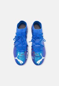Scarpe da calcio blu con finitura texture, accenti rossi dinamici, caviglie imbottite e un logo bianco prominente sul lato. I lacci sono centrali.