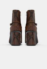 Botas de tobillo marrones de tacón con textura de piel de serpiente, que cuentan con una cremallera trasera y un tacón ancho. Correas con hebillas acentúan el diseño.