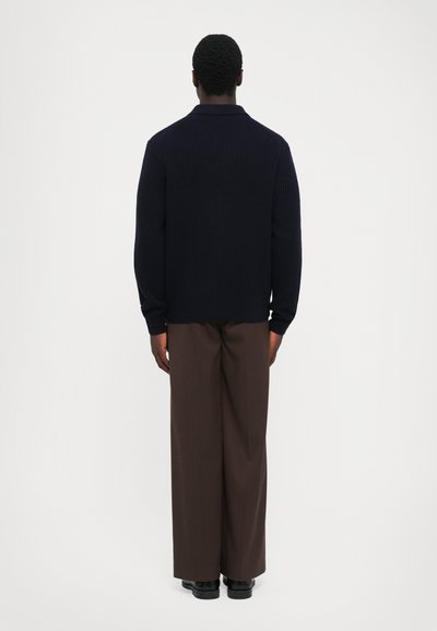 Hombre de pie, de espaldas, vistiendo un suéter negro de canalé, pantalones anchos marrones y zapatos negros, contra un fondo blanco liso.