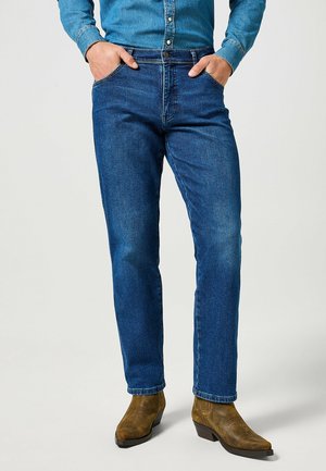 Homme portant un jean denim bleu, une chemise bleue boutonnée, les mains dans les poches, et des bottes de cowboy en cuir marron, debout sur un fond clair.