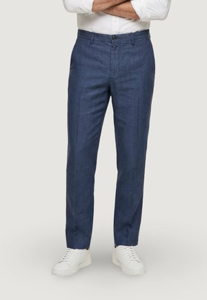 Uomo che indossa pantaloni blu su misura, scarpe da ginnastica bianche e una camicia bianca a maniche lunghe con le braccia incrociate, in piedi su uno sfondo semplice.