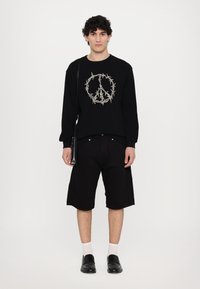 Black sweatshirt with peace symbol embroidered in beige, paired with black knee-length shorts and black loafers. Model holds a small black bag.