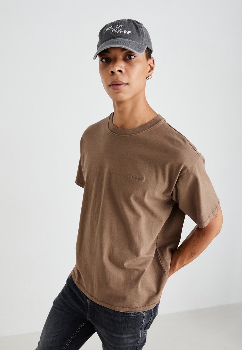 Braunes Baumwoll-T-Shirt mit Rundhalsausschnitt und kurzen Ärmeln, mit einem dezenten Logo auf der Brust. Kombiniert mit einer grauen Kappe, die Text zeigt.