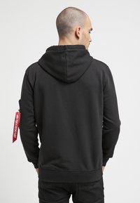 Sudadera negra con capucha, que muestra una textura suave. Presenta un bolsillo con cremallera lateral y una etiqueta de tela roja en la manga.