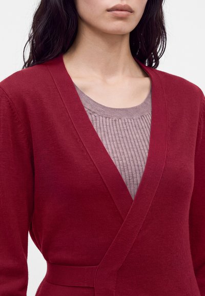 Femme portant un cardigan cache-cœur rouge profond sur un haut côtelé violet clair, montré du cou à la taille sur fond blanc.