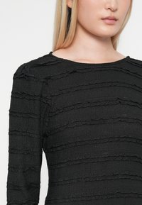 Vestido preto texturizado com padrões de babados horizontais, mangas longas e decote redondo. O material parece pesado e em camadas.