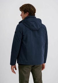 Man met bruin haar, gekleed in een marineblauwe hoodie en olijfgroene broek, met zijn rug naar de camera tegen een effen lichte achtergrond.