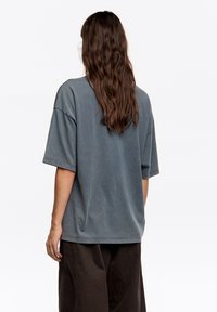 Szara, oversize bawełniana koszulka z opuszczonymi ramionami i krótkimi rękawami, o luźnym kroju i miękkiej fakturze, pokazana od tyłu.