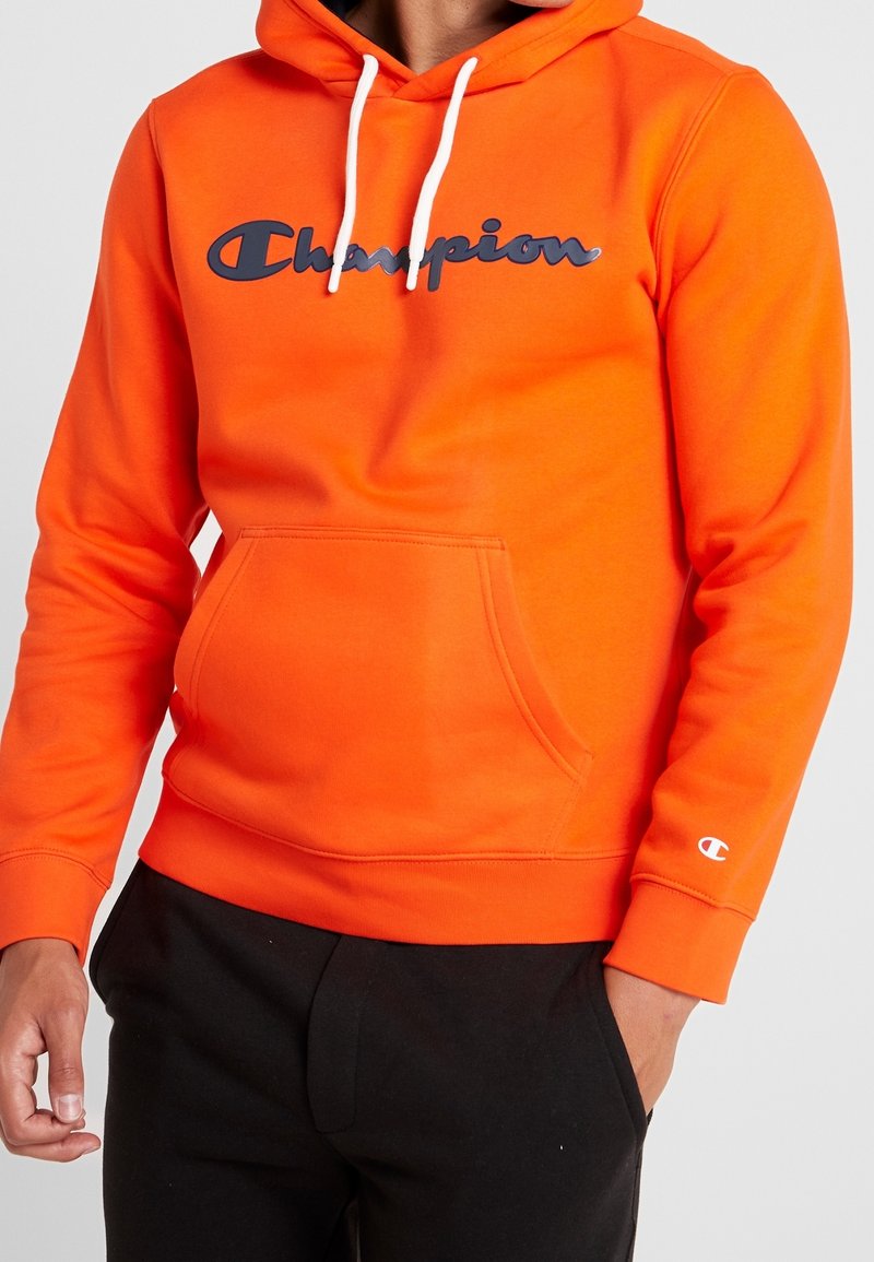 Sudadera naranja hecha de tejido suave, con un bolsillo tipo canguro y un gran logo de "Champion" en azul marino en el frente. Cuerdas blancas presentes.