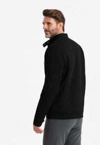 Veste noire à col haut, en tissu texturé, avec des détails de couture visibles et des fermetures éclair. Portée sur un pantalon gris, mettant en valeur une coupe moderne.