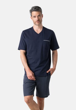 Homme aux cheveux courts gris portant un T-shirt bleu marine à col en V avec une poche poitrine et un short bleu marine à motifs, debout avec une main dans la poche.