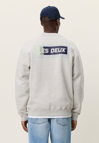 Άνδρας που φοράει ανοιχτό γκρι φούτερ με το λογότυπο «LES DEUX SPORTING GOODS» στην πλάτη, μπλε καπέλο και γαλάζια τζιν, γυρισμένος με την πλάτη στην κάμερα.