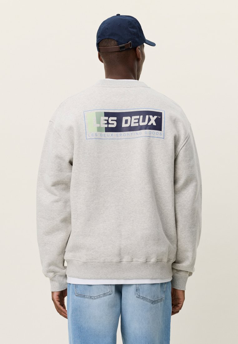 Άνδρας που φοράει ανοιχτό γκρι φούτερ με το λογότυπο «LES DEUX SPORTING GOODS» στην πλάτη, μπλε καπέλο και γαλάζια τζιν, γυρισμένος με την πλάτη στην κάμερα.