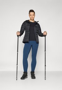 Veste noire rembourrée, haut sombre, leggings bleus et bottes de randonnée noires. Tient des bâtons de trekking, debout sur un fond blanc.