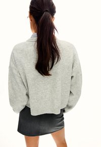 Kvinna med långt mörkt hår i hästsvans, iförd en ljusgrå oversized tröja och en kort svart kjol, vänd bort från kameran.