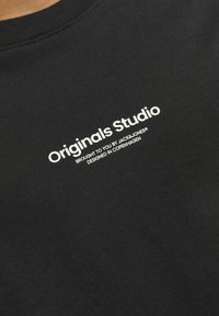 Camiseta de algodón negra con cuello redondo acanalado, que presenta el texto impreso en blanco "Originals Studio" y detalles de marca. Textura suave.