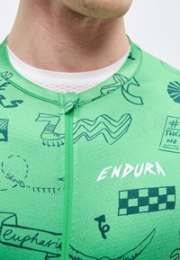 Κοντινό πλάνο ενός ατόμου που φοράει πράσινο ποδηλατικό μπλουζάκι με μαύρα σχέδια και τη λέξη "ENDURA" τυπωμένη κοντά στο φερμουάρ.