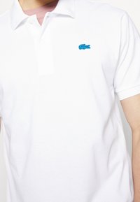 Polo shirt blanc en coton avec un col, présentant un logo de crocodile brodé bleu sur la poitrine gauche. Manches courtes, coupe classique.