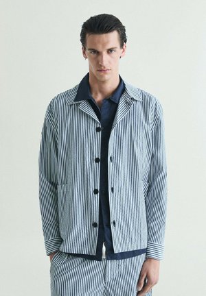 Joven con chaqueta y pantalones a rayas verticales a juego y una camisa oscura debajo, de pie frente a un fondo liso.
