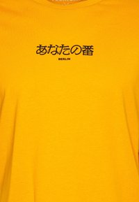 Camiseta de algodón amarilla con texto negro en japonés e inglés que dice "あなたの番" y "BERLÍN". Textura suave con diseño de cuello redondo.