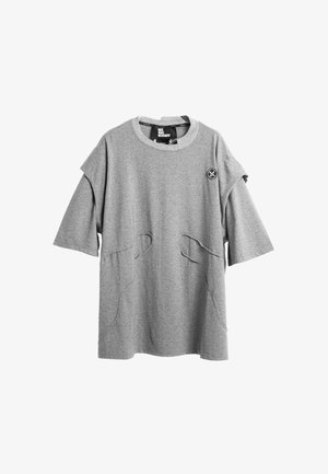Camiseta gris de gran tamaño con diseño cuadrado y dos bolsillos frontales, combinada con pantalones grises que presentan un detalle de cadena lateral y puños elásticos.