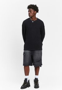 Svart langermet genser i strikk, grå cargo shorts med flere lommer, hvite sokker og svarte støvler. Enkel design og avslappet passform.