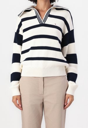 Femme portant un pull blanc avec de larges rayures horizontales bleu marine et un pantalon beige, debout devant un fond blanc uni.