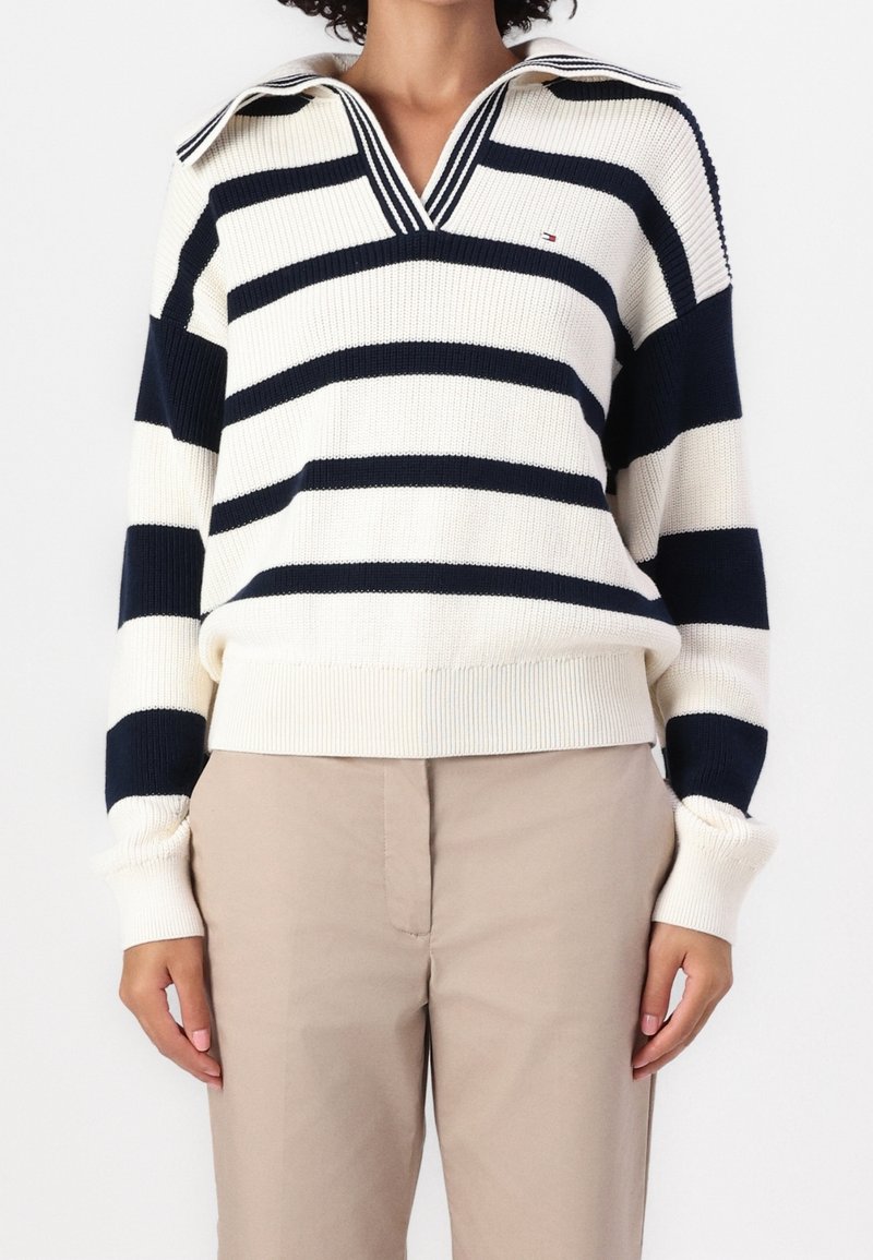 Femme portant un pull blanc avec de larges rayures horizontales bleu marine et un pantalon beige, debout devant un fond blanc uni.