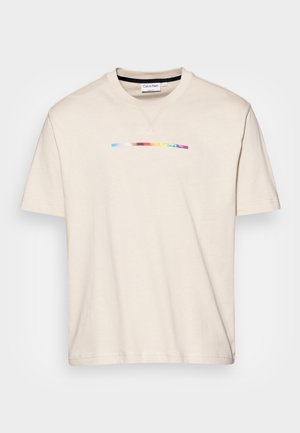 Beżowa koszulka Calvin Klein z krótkim rękawem, okrągłym dekoltem i cienkim poziomym paskiem w kolorach tęczy na wysokości klatki piersiowej.