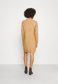 Donna con capelli ricci che indossa un vestito di maglione color cammello lungo fino al ginocchio e stivaletti marroni, girata di spalle su uno sfondo bianco semplice.