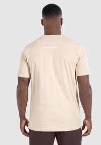 Beige bomulds T-shirt med korte ærmer og rund hals, prydet med "SMILODOX" i hvid tekst på den øverste del af ryggen. Glat, blød tekstur.