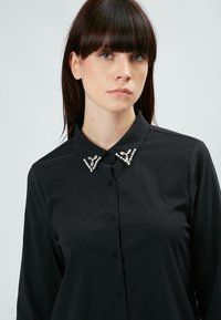 Camisa negra de manga larga con botones en el frente, que presenta un collar adornado con acentos de cuentas plateadas en un patrón decorativo triangular.