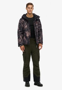 Gewatteerde jas in zwart met roze gemarmerd patroon, voorzien van een capuchon en een ritssluiting aan de voorkant; gecombineerd met olijfgroene cargobroeken en zwarte laarzen.