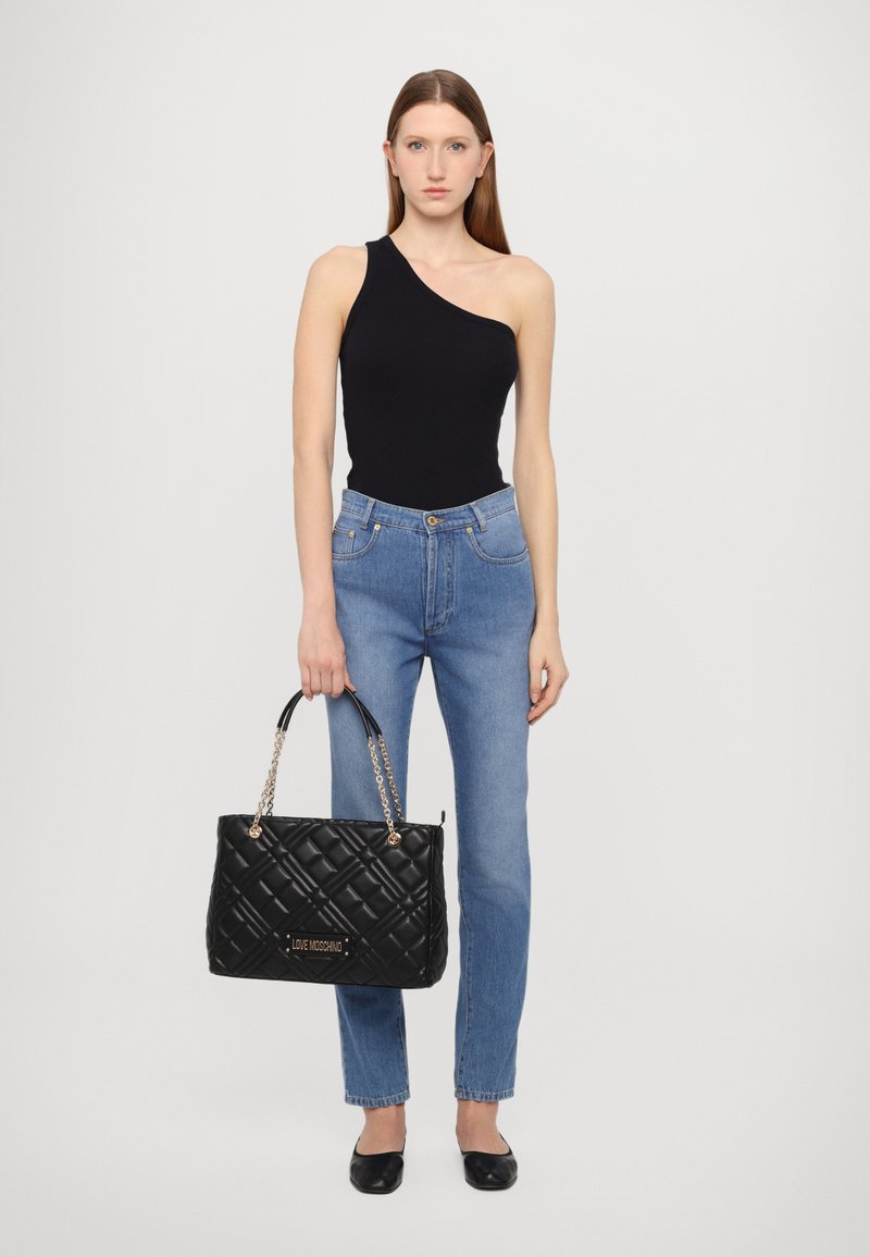 Borsa a tracolla nera trapuntata con dettagli in oro, doppi manici a catena e in pelle. Modello che indossa un top nero a spalla unica e jeans azzurri chiari.