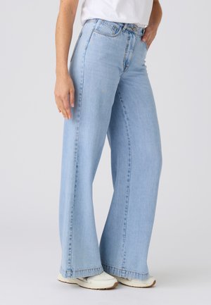 Persoon draagt lichtblauwe jeans met wijde pijpen, witte sneakers en een wit shirt, staand met één hand in de zak en voeten iets uit elkaar.