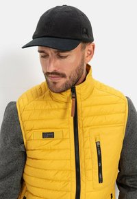 Gul puffervest med et gråt langærmet lag under, der har lynlåslommer, quiltet tekstur og en sort kasket. Jakken har lynlåslukning.