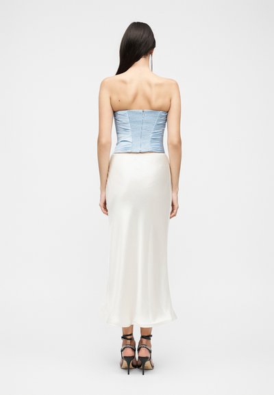 Γυναίκα με μακριά σκουρόχρωμα μαλλιά, που φοράει ανοιχτό μπλε κορσέ χωρίς τιράντες, λευκή σατέν μεσάτη φούστα και μαύρα σανδάλια με ψηλό τακούνι, στέκεται με την πλάτη προς τα πίσω.