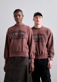 Brun sweatshirt med sort "Aries" grafik og print på ærmerne. Model iført en sort nederdel, anden model i mørke cargo-bukser, begge i afslappet pasform.