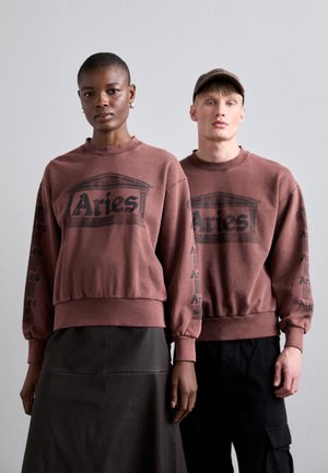 Brun genser med svart "Aries" grafikk og trykk på ermene. Modell iført et svart skjørt, den andre modellen i mørke cargos, begge i en avslappet passform.