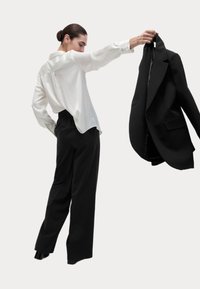 Weißes Satinhemd mit lockerer Passform, schwarze weit geschnittene Hose und schwarzer Blazer, der in einer Hand gehalten wird. Glatte Textur, klassisches Design.