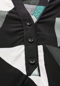 Tissu noir avec des motifs géométriques en blanc, teal et gris. Présente une patte de boutonnage avec quatre boutons noirs. Texture lisse.