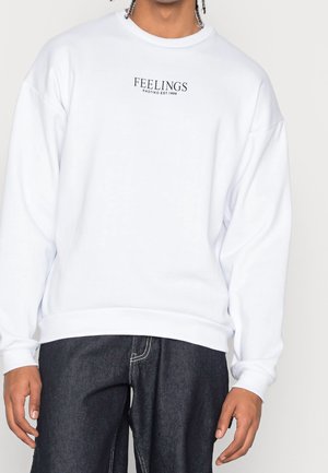 Sweatshirt blanc à col rond et épaules tombantes. Présente un texte noir "FEELINGS" sur la poitrine, avec des poignets et un ourlet côtelés.