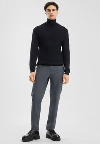 Čierny rolák, slim fit, v kombinácii so šedými nohavicami. Model má na sebe čierne šnurovacie topánky s hrubou podrážkou a stojí pred jednoduchým pozadím.