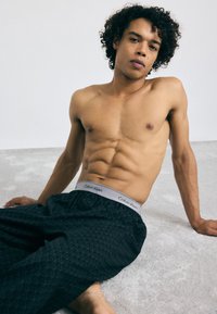 Čierne nohavice s vzorom na pohodlné nosenie s šedým pásom Calvin Klein. Model je bez trička, ukazuje vytrénovanú postavu a sedí na svetlom povrchu.
